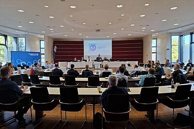 Menschen sitzen in einem Saal. Im Hintergrund ist eine Empore mit einer Sitzreihe. Dazwischen steht ein Mann und hält eine Rede.